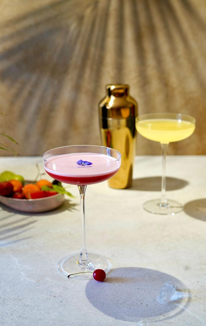 A pink cocktail and a pale cocktail next to a brass shaker